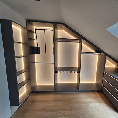 In der Mitte des Bildes ein offener, begehbarer Einbauschrank in der Dachschräge (Giebelwand) aus Dekorplatte (Achatgrau). Ausstattung: Praktische Kleiderstangen, Kleiderlift, Hosenauszug und Fachböden. Außerdem links ein Wandregal und rechts eine Kommode. Rückwände aus weißem Acrylglas, mit LED hinterleuchtet.