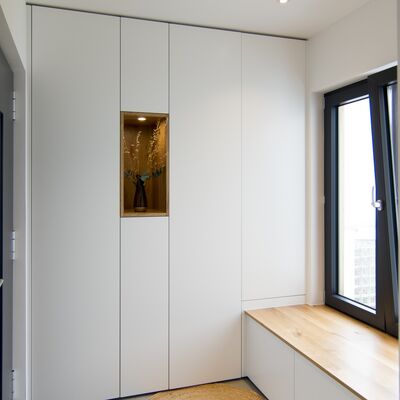 Großer, eleganter Garderobenschrank mit weißen Fronten und einem offenen Dekofach in der Mitte aus Eiche. Rechts schließt eine Sitzbank mit Stauraum in gleichem Stil an.